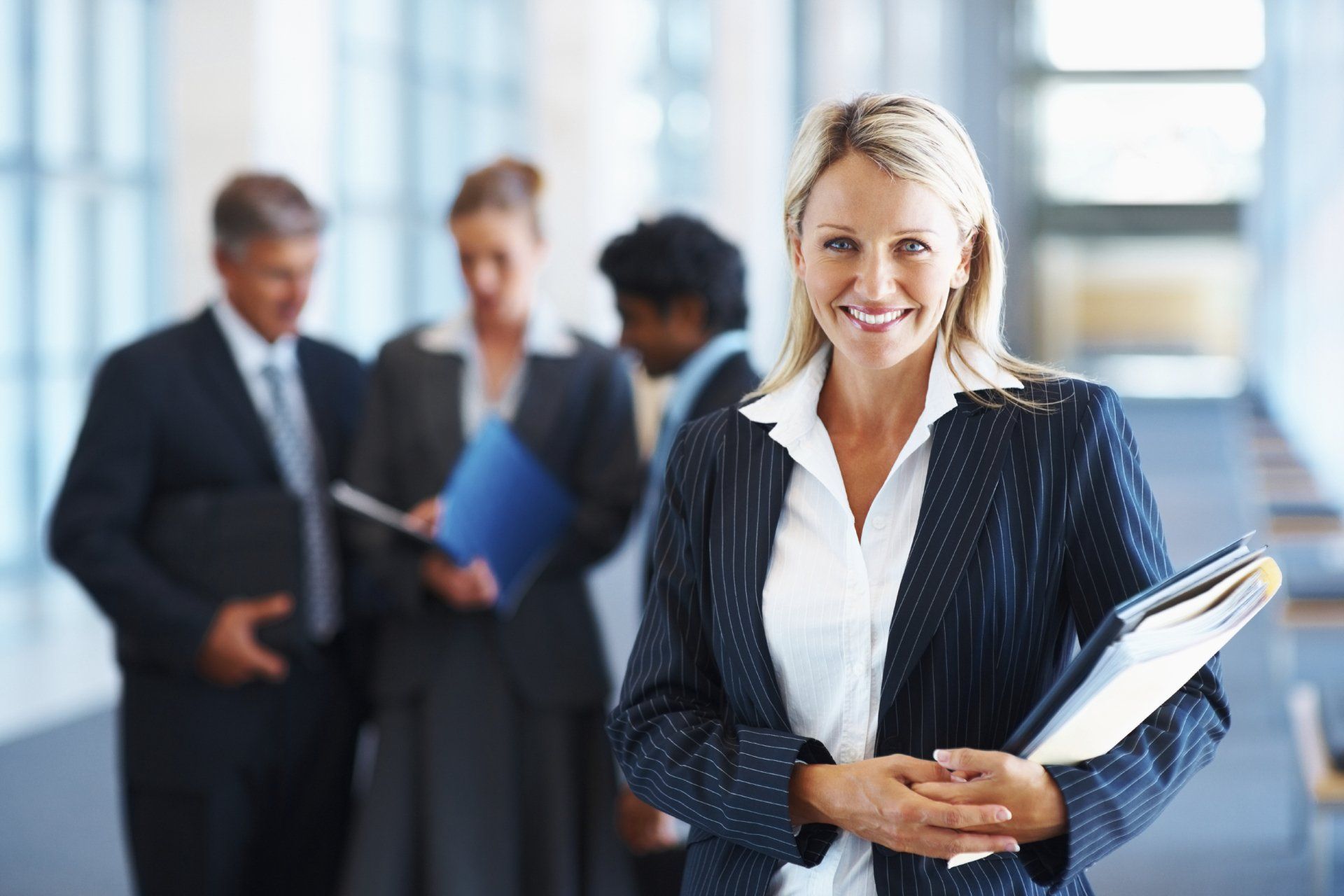 A smiling professional in a pinstriped blazer holds documents in a bright, modern office with colleagues in the background.