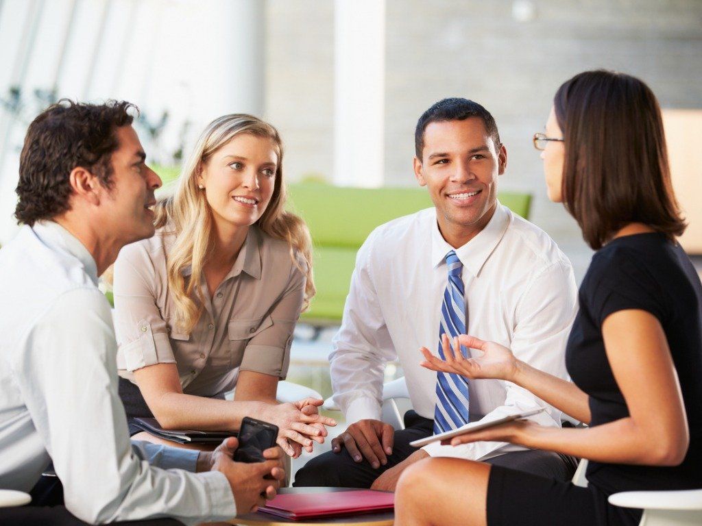Four professionals in business attire sit in a circle in an office, actively discussing work while holding documents.