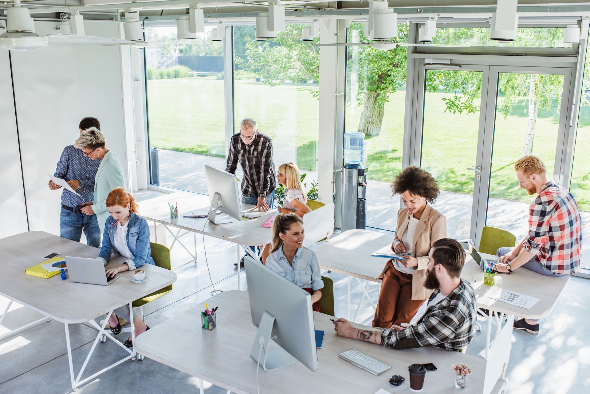 A diverse team collaborates in a bright, modern office with large windows, working at desks with computers and notebooks.