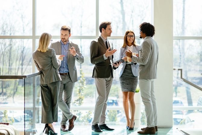 Five professionals in business attire stand in a sunlit office lobby, conversing and holding folders.