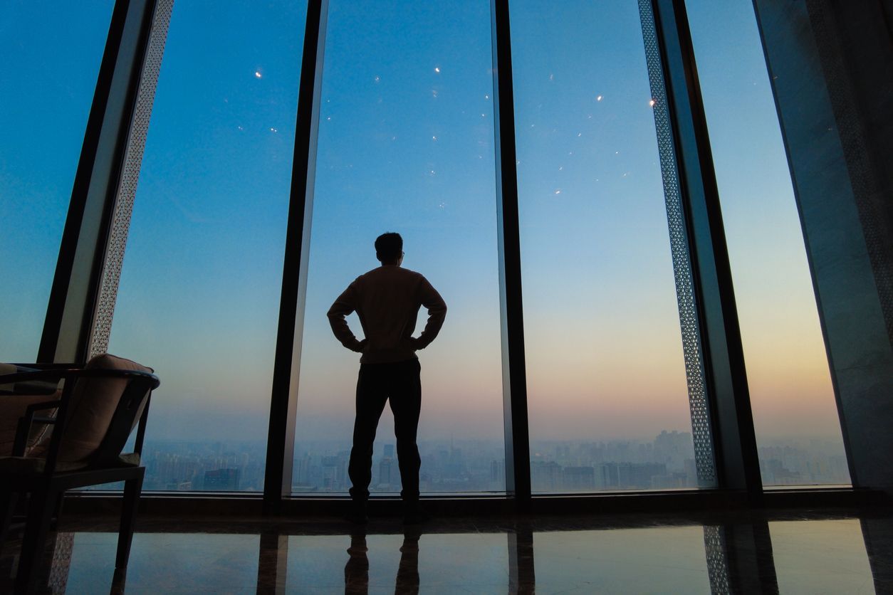 A silhouette of a person standing with hands on hips, looking out of a large floor-to-ceiling window at a city at dusk.