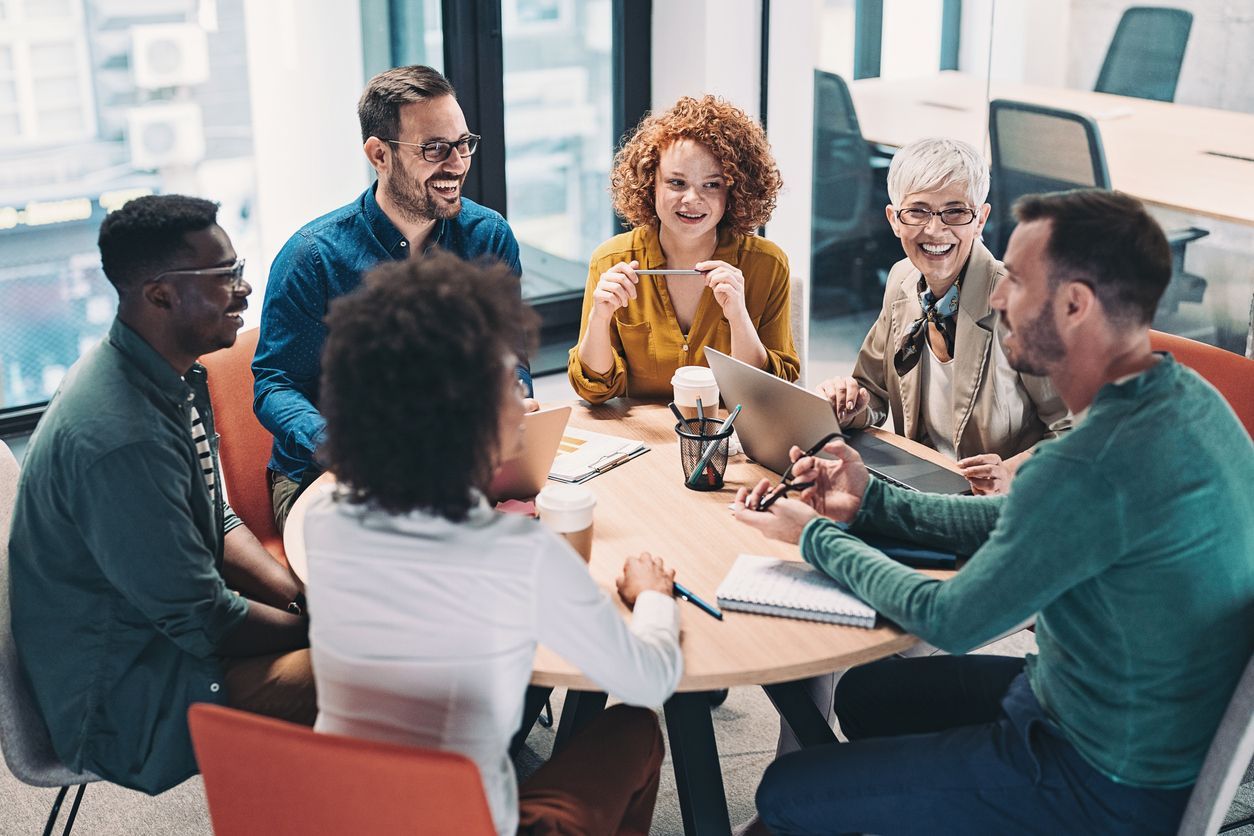 A group of colleagues sitting around a circular table in an office, laughing and collaborating during a meeting.