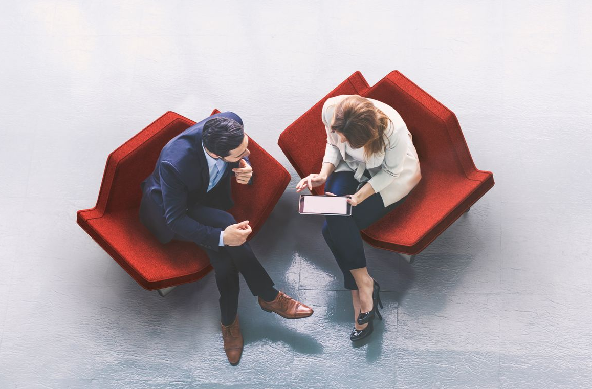 A high-angle view of two people in business attire sitting in red armchairs, conversing while looking at a tablet.