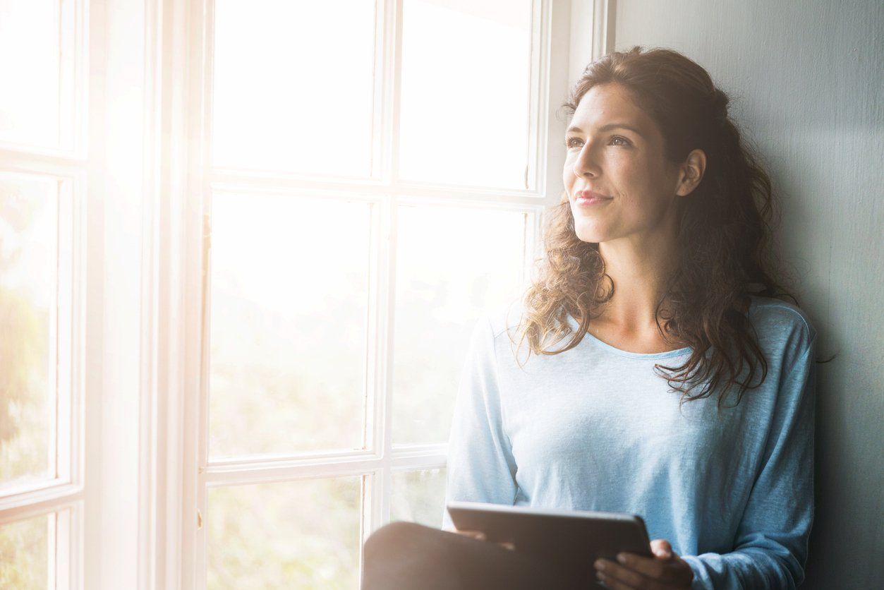 A person with wavy hair sitting by a window, holding a tablet and looking out with a gentle smile in soft, bright light.