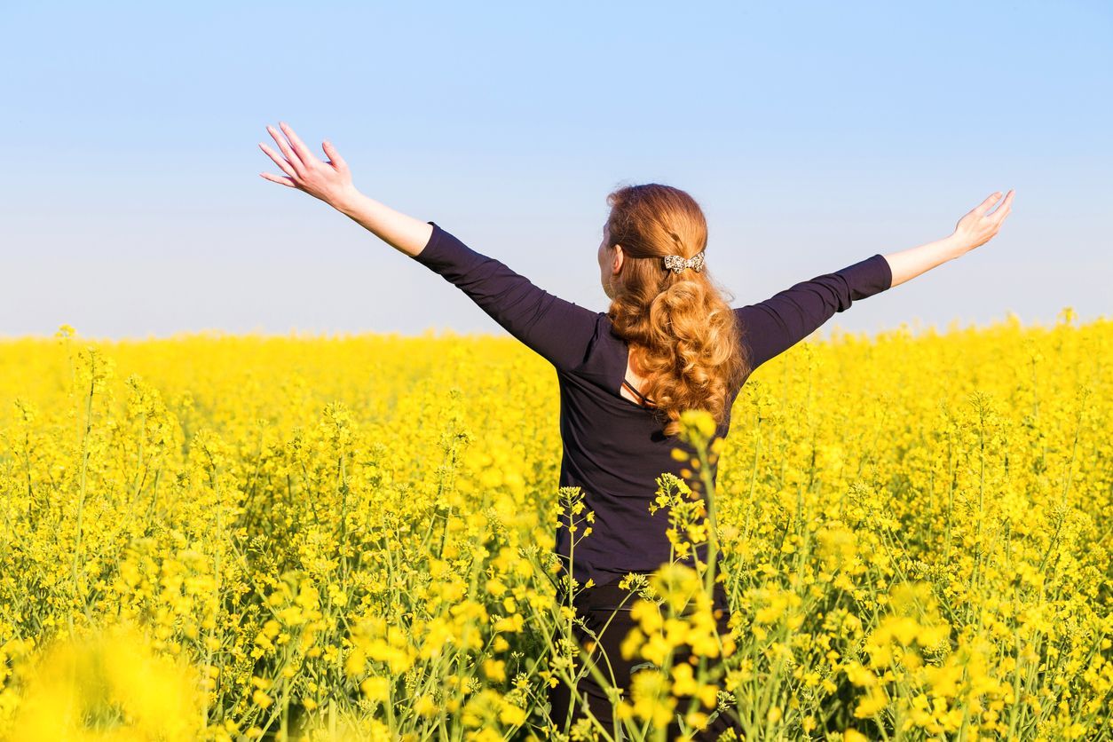 A person standing in a bright yellow field of flowers with arms outstretched toward a clear blue sky.
