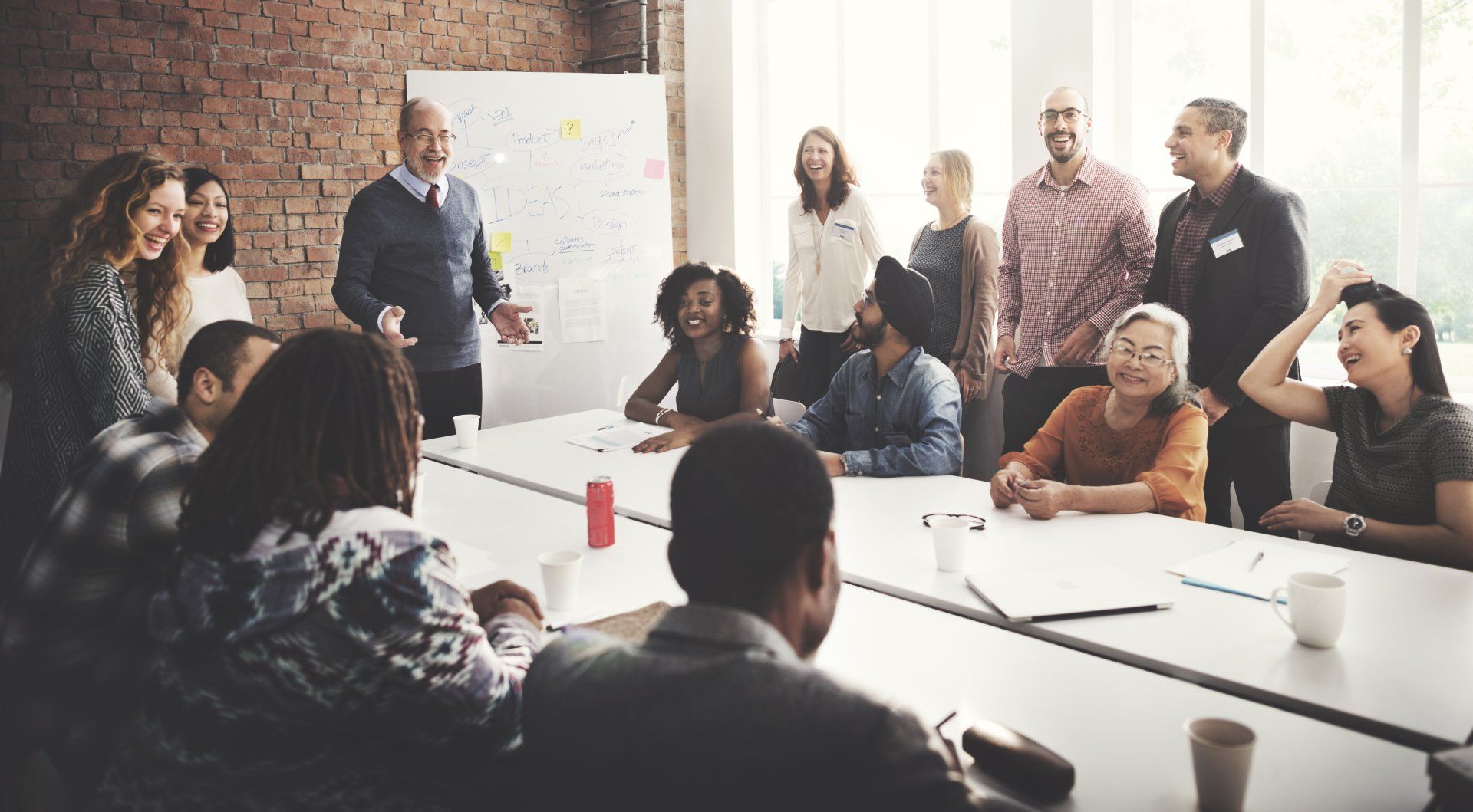 A diverse group of people in a bright office meeting room listen to a man presenting by a whiteboard.