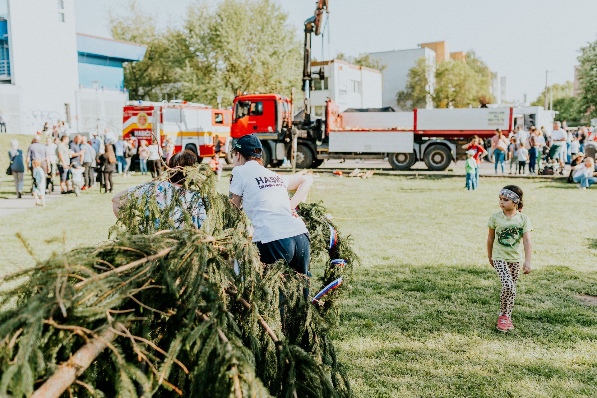 Children and adults at a park beside a red truck, with one child carrying branches on grass