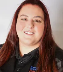 Smiling person with long reddish hair wearing a black collared shirt against a light background