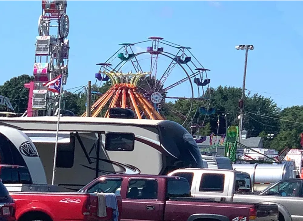 Amusement park midway with ferris wheel and ride behind parked cars and buses on a sunny day