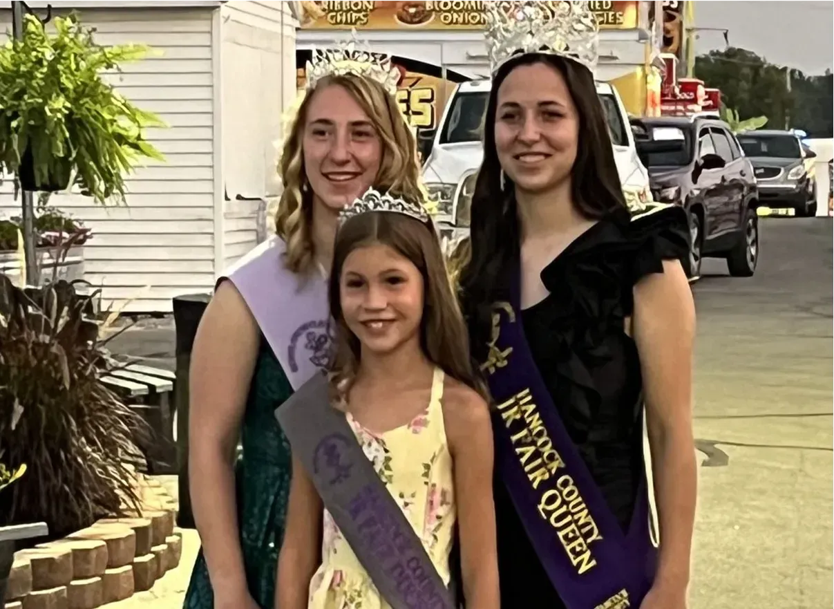 Three smiling sash-wearing pageant winners pose outdoors near a building and parked cars.
