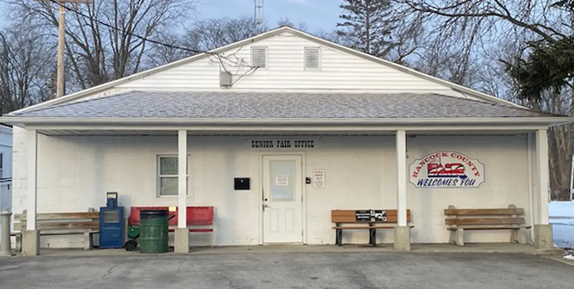 Small white building with a front porch, benches, trash bins, and a sign on the right side.