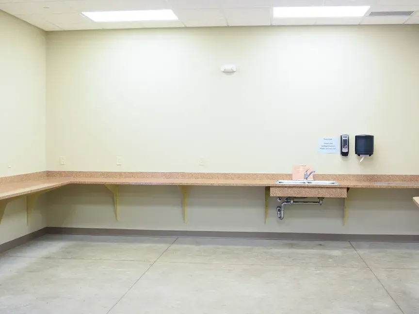Empty indoor room with beige walls, long counter, sink, and wall-mounted dispensers