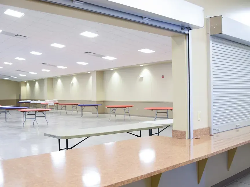 Empty cafeteria with long tables and benches under bright ceiling lights, viewed from a service counter.
