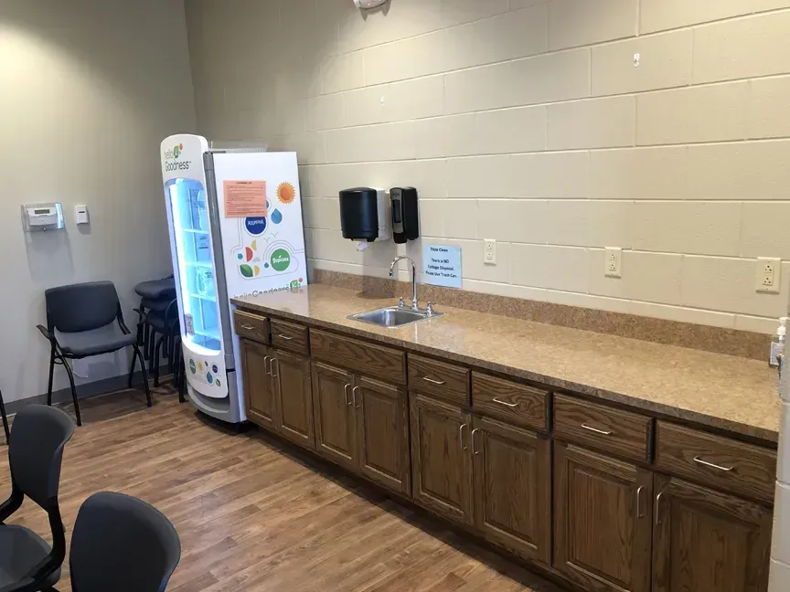 Break room kitchenette with sink, cabinets, water cooler, vending machine, and chairs on wood flooring