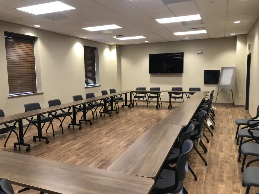 Empty conference room with U-shaped tables, black chairs, wall-mounted TV, and whiteboard.