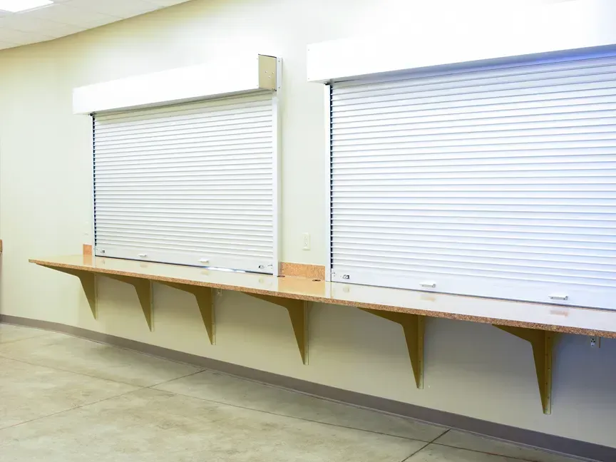 Empty indoor service counter with two closed white roll-up shutters and wooden shelves underneath