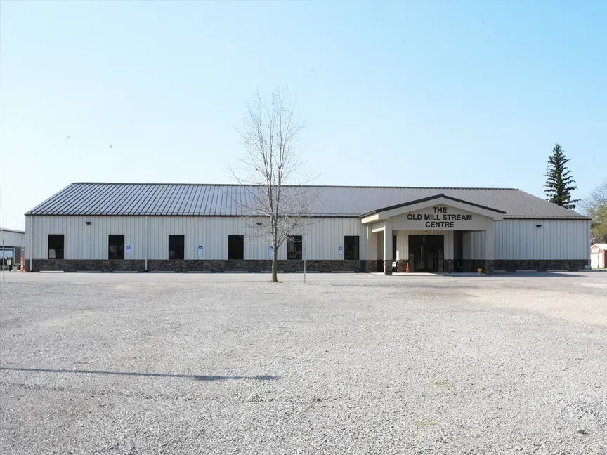 Single-story white building with a metal roof and central entrance, viewed across a gravel lot