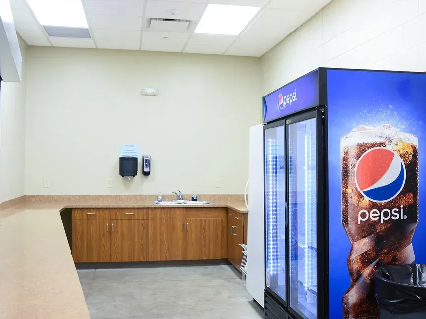 Empty break room with wooden counter, sink, and Pepsi vending cooler against a beige wall
