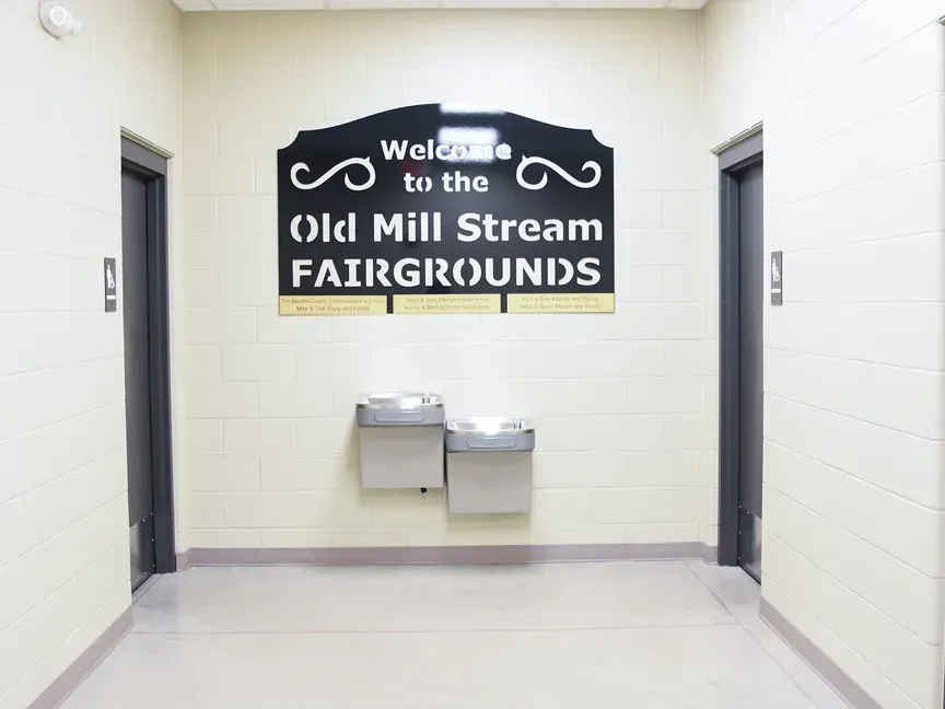 Hallway sign reading โWelcome to the Old Mill Stream Fairgroundsโ above two water fountains