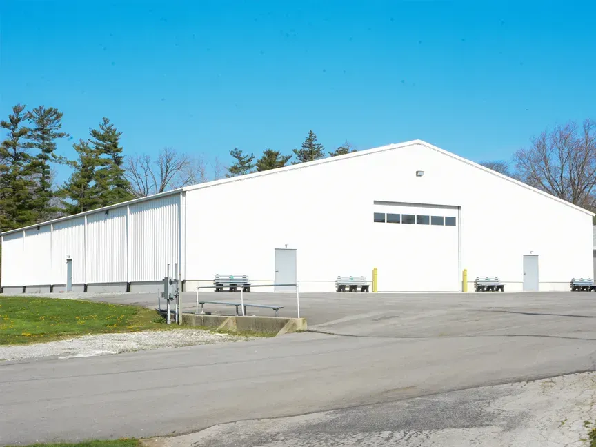 Large white warehouse building with a long side wall, parked trucks, and a paved lot under a clear blue sky