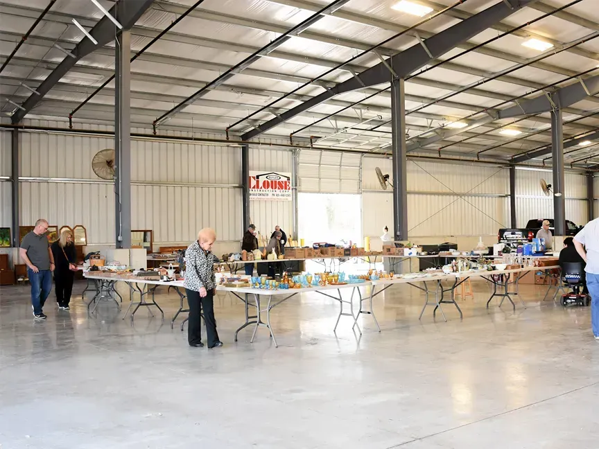 Large indoor warehouse hall with folding tables set up for an event and a few people standing around