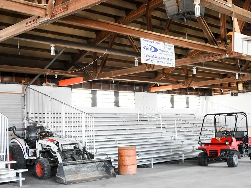 Sheltered storage area with tractors, a utility vehicle, and stacked metal bleachers under a wooden roof