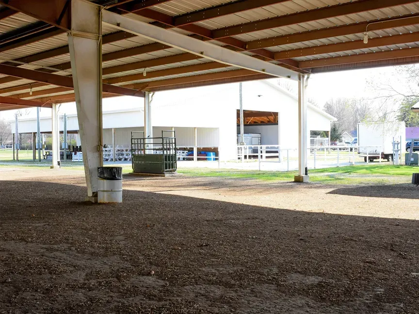 Covered outdoor pavilion with wooden roof, concrete pillars, and a gravel floor beside a fenced park area.