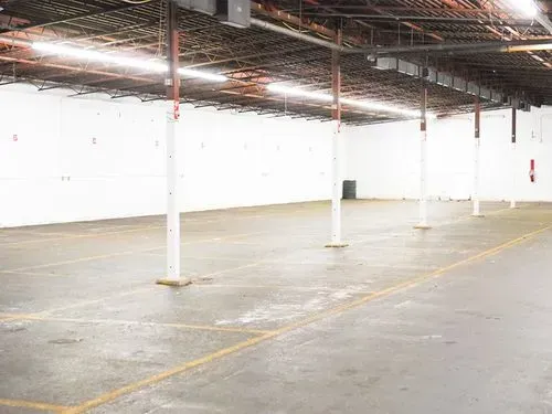 Empty warehouse with white support columns and concrete floor, lit by overhead industrial lights