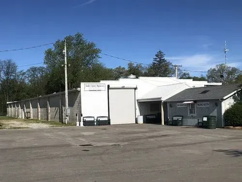 Small white building with a dumpster and fence beside an empty paved lot under a blue sky