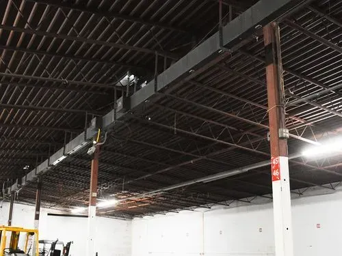 Empty warehouse interior with exposed black ceiling beams and white walls, lit by bright overhead lights