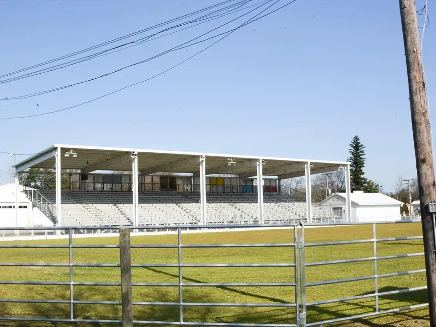 Empty outdoor stadium with white stands, green field, and a metal fence in the foreground