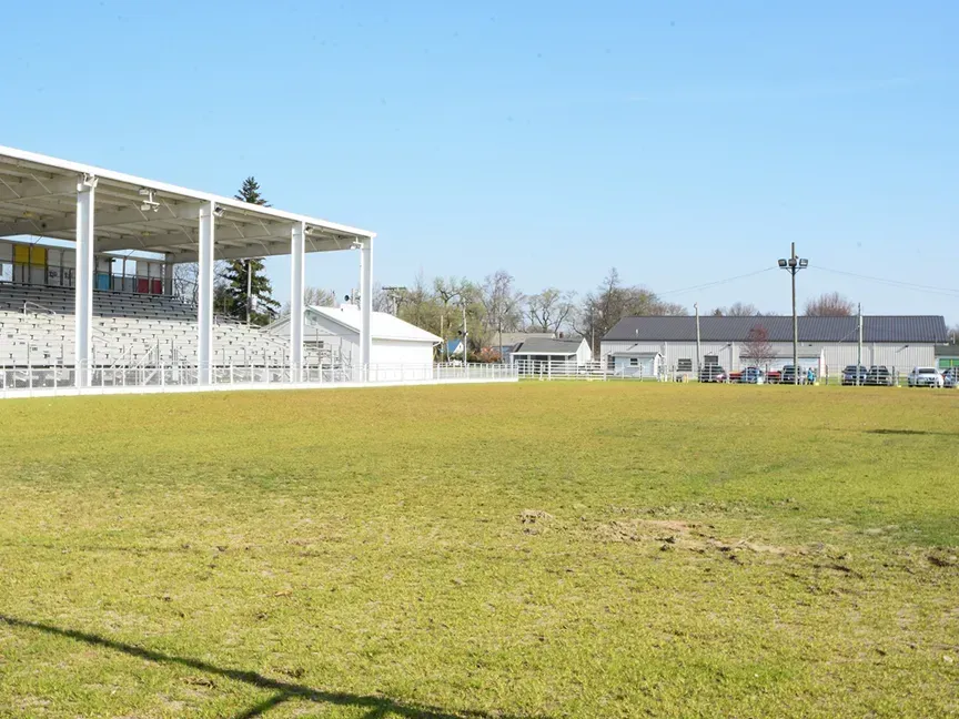 Empty grassy sports field beside white bleachers and a low stadium building under a clear blue sky