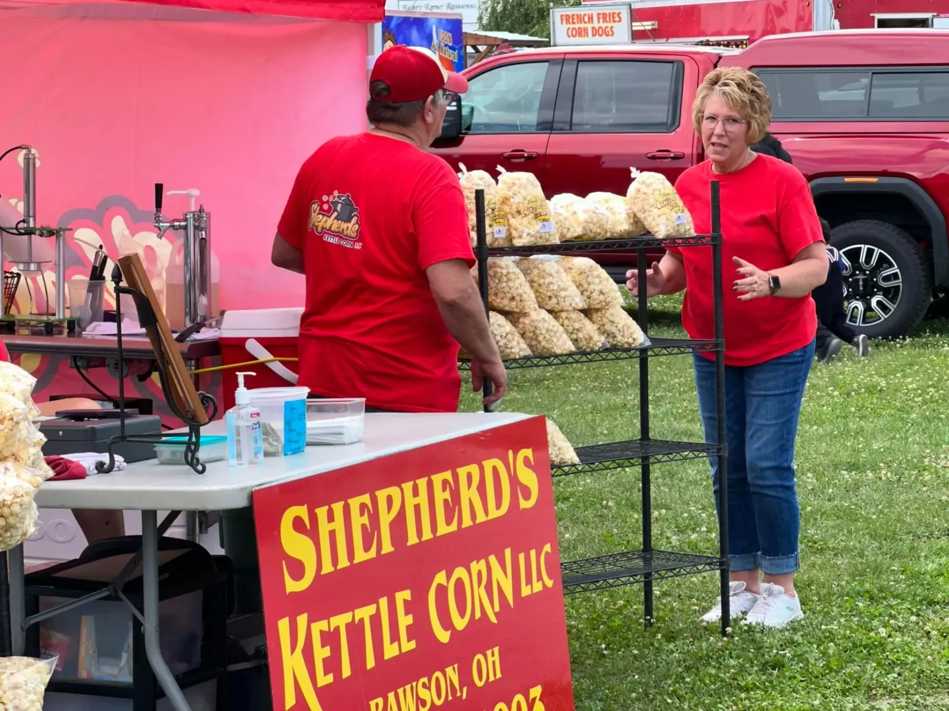 Two people in red shirts at a Shepherd’s Kettle Corn booth with bags of popcorn and a red truck behind them
