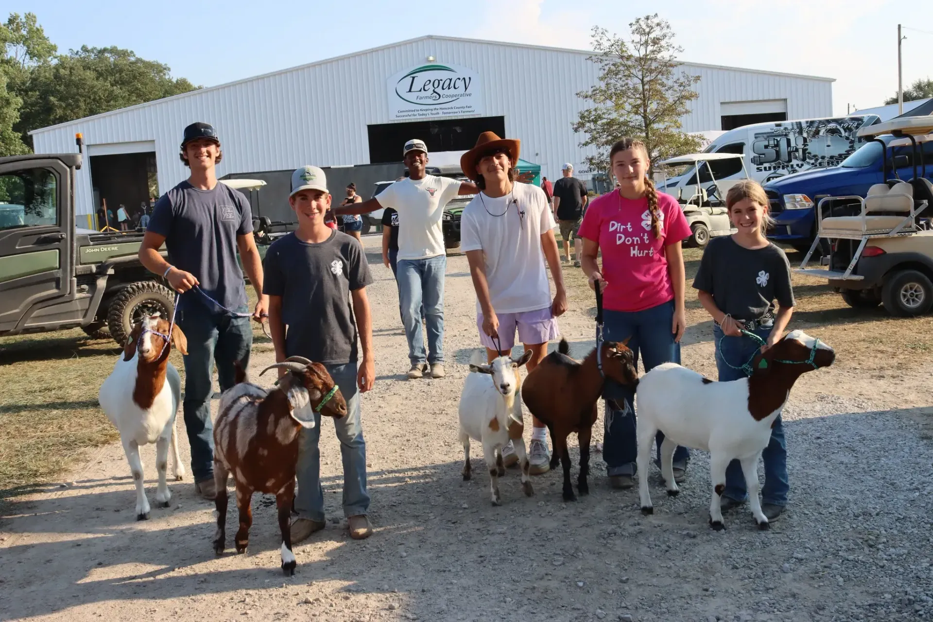 People holding goats and calves in front of a white barn and parked trucks outdoors