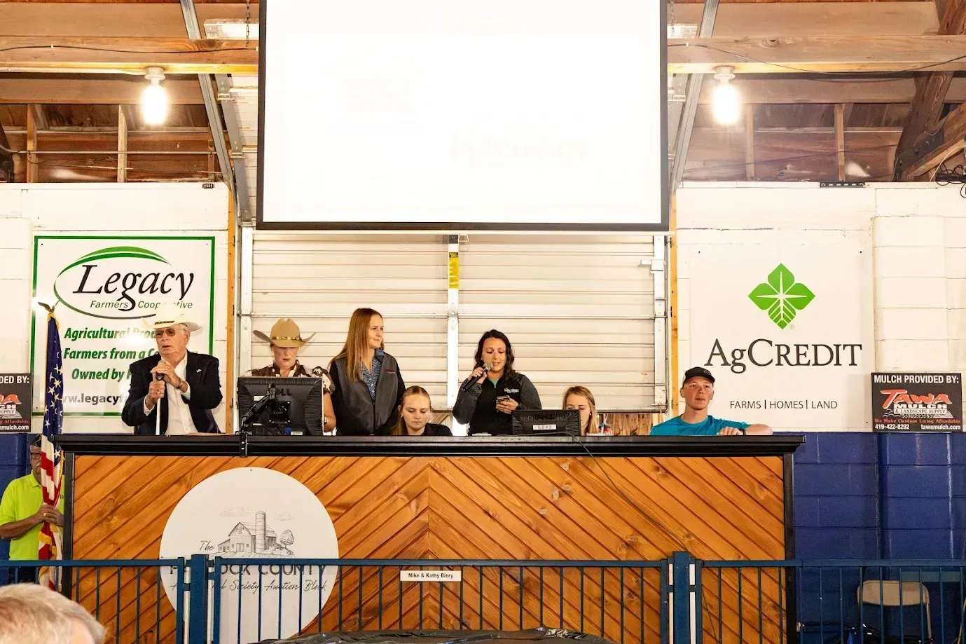 People at a long table on a stage in a barn, with Legacy and AgCredit signs behind them.