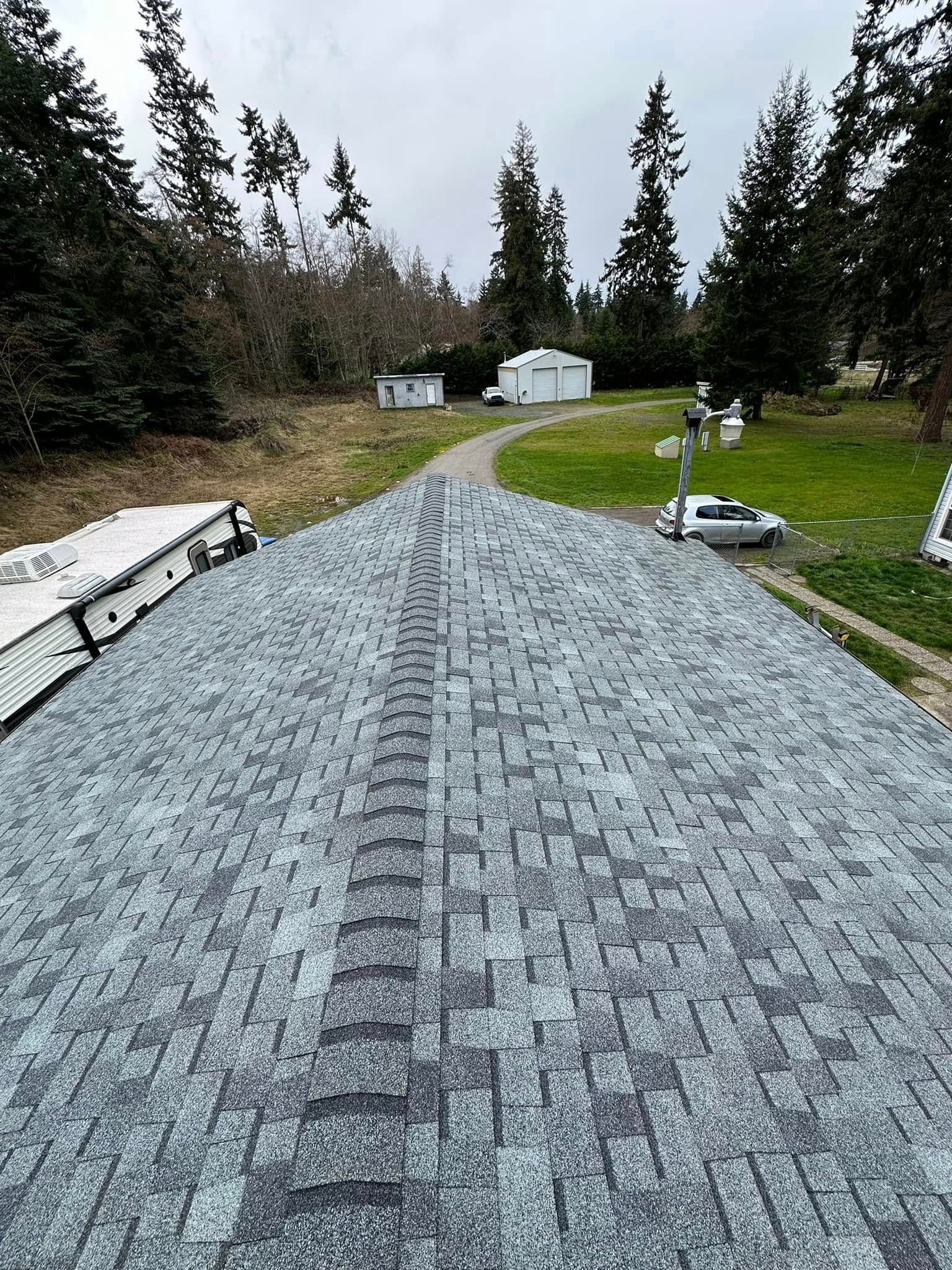Newly shingled roof, gray color, with surrounding trees and a small road leading to buildings in the distance.