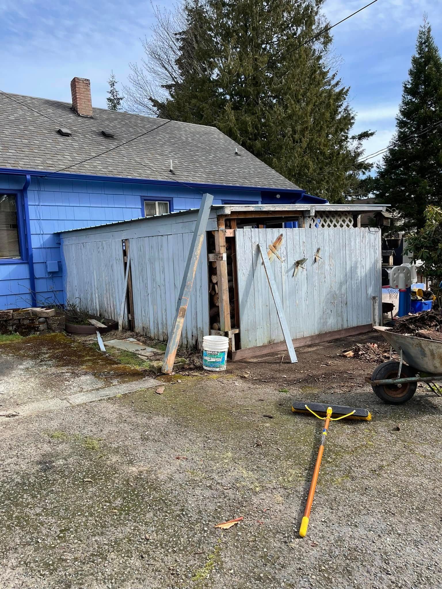 Blue house with deteriorating wooden structure; construction materials scattered on gravel ground.