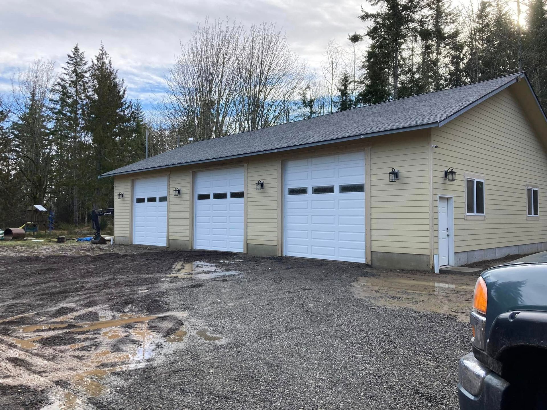Yellow garage with three white garage doors, gravel driveway, and trees.