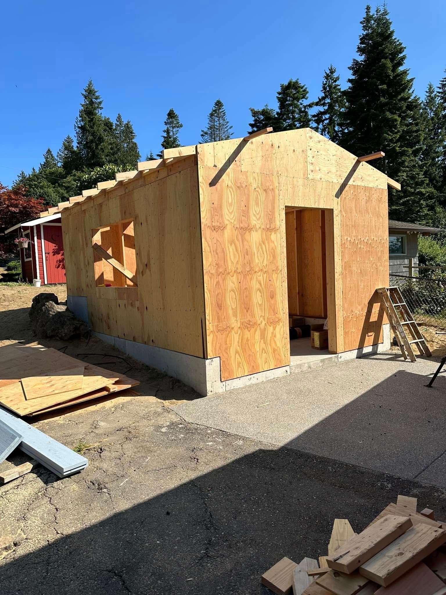 Wooden shed in progress on a gravel base, with a door and window openings, under a clear, blue sky.