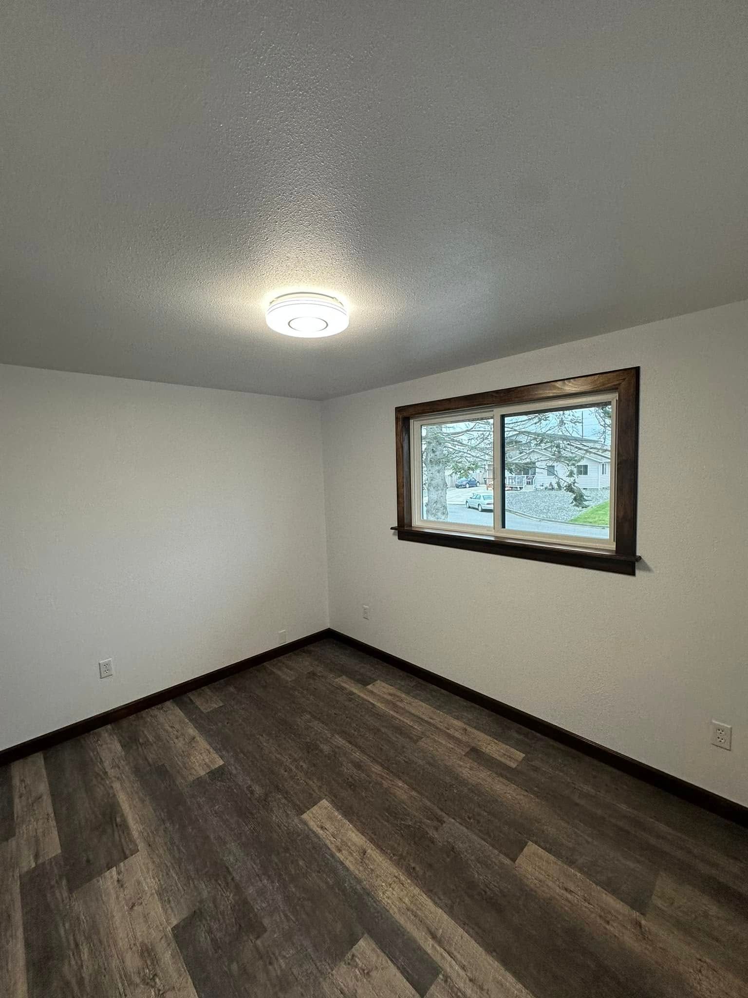 Empty room with wood-look flooring, white walls, a window with a brown frame, and a ceiling light.