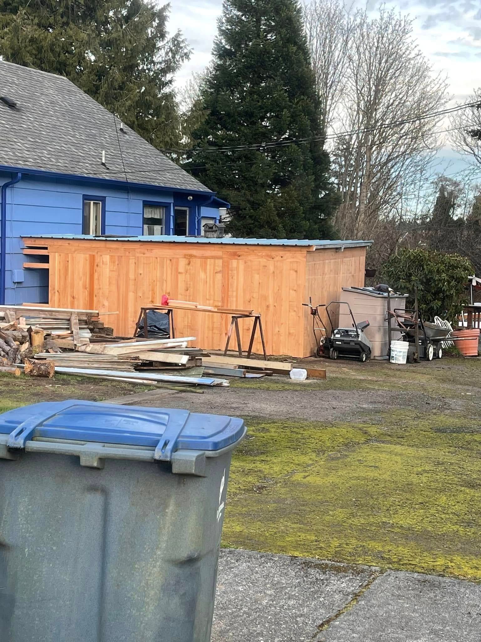 Blue-sided house under construction, shed, trash can, and yard debris.
