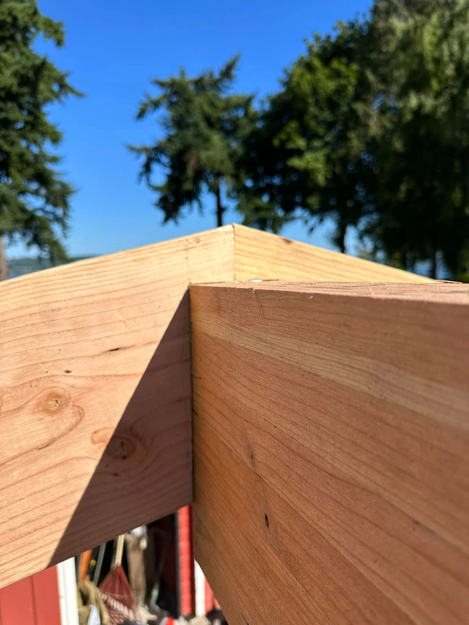 Close-up of a wooden structure's corner, showcasing angled cuts meeting at a peak, set against a blue sky.