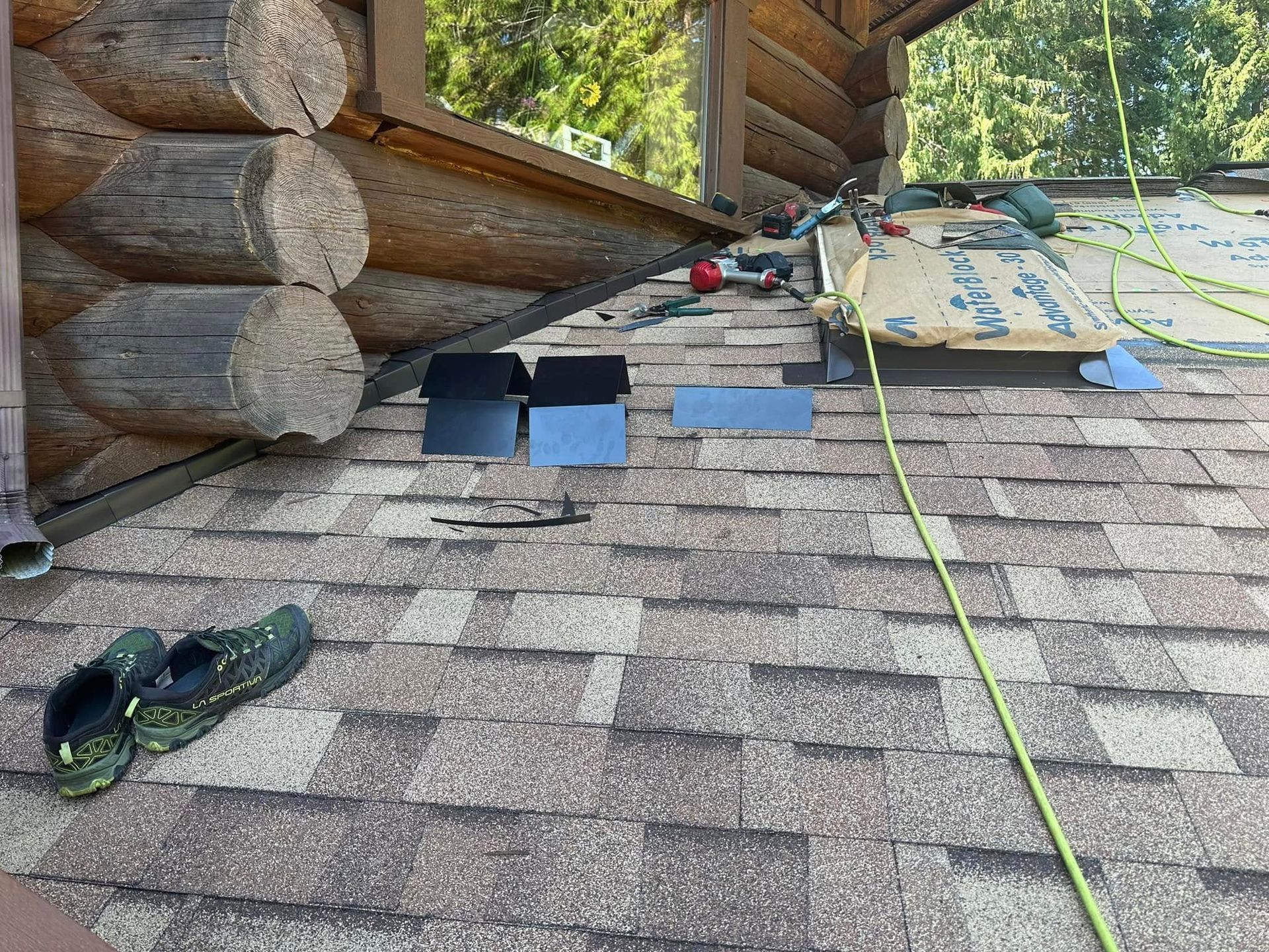 Roof with shingle samples, tools, and work boots on top. Log cabin and trees in background.