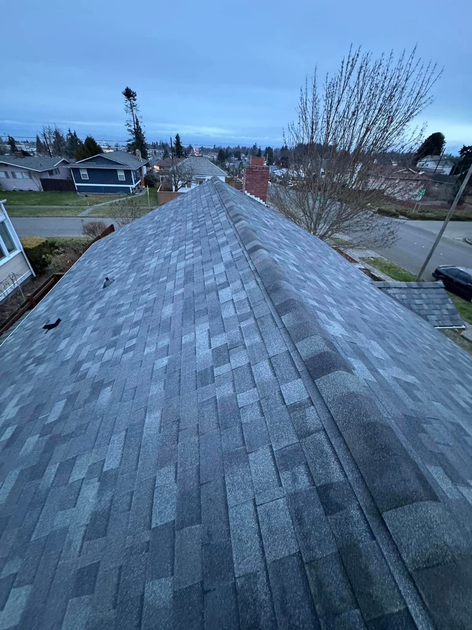 View of a gray asphalt shingle roof on a house, street, trees, and sky in the background.