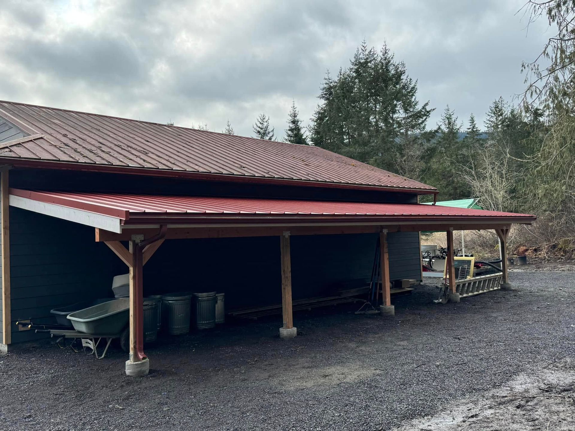 A red roofed carport attached to a dark building. Gray gravel ground and overcast sky.