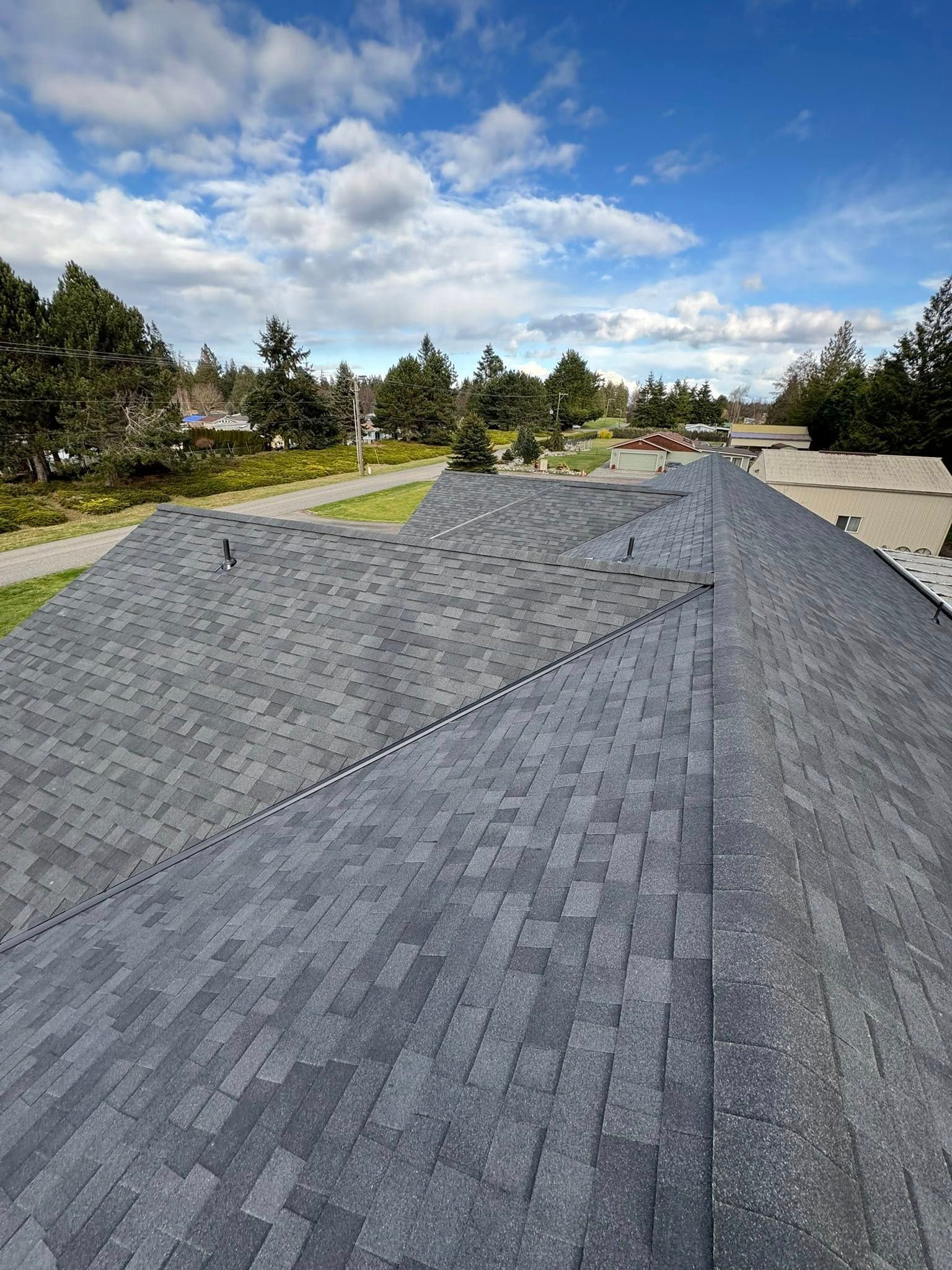 Gray asphalt shingle roof against a blue sky with fluffy clouds; trees and a pathway visible in the distance.