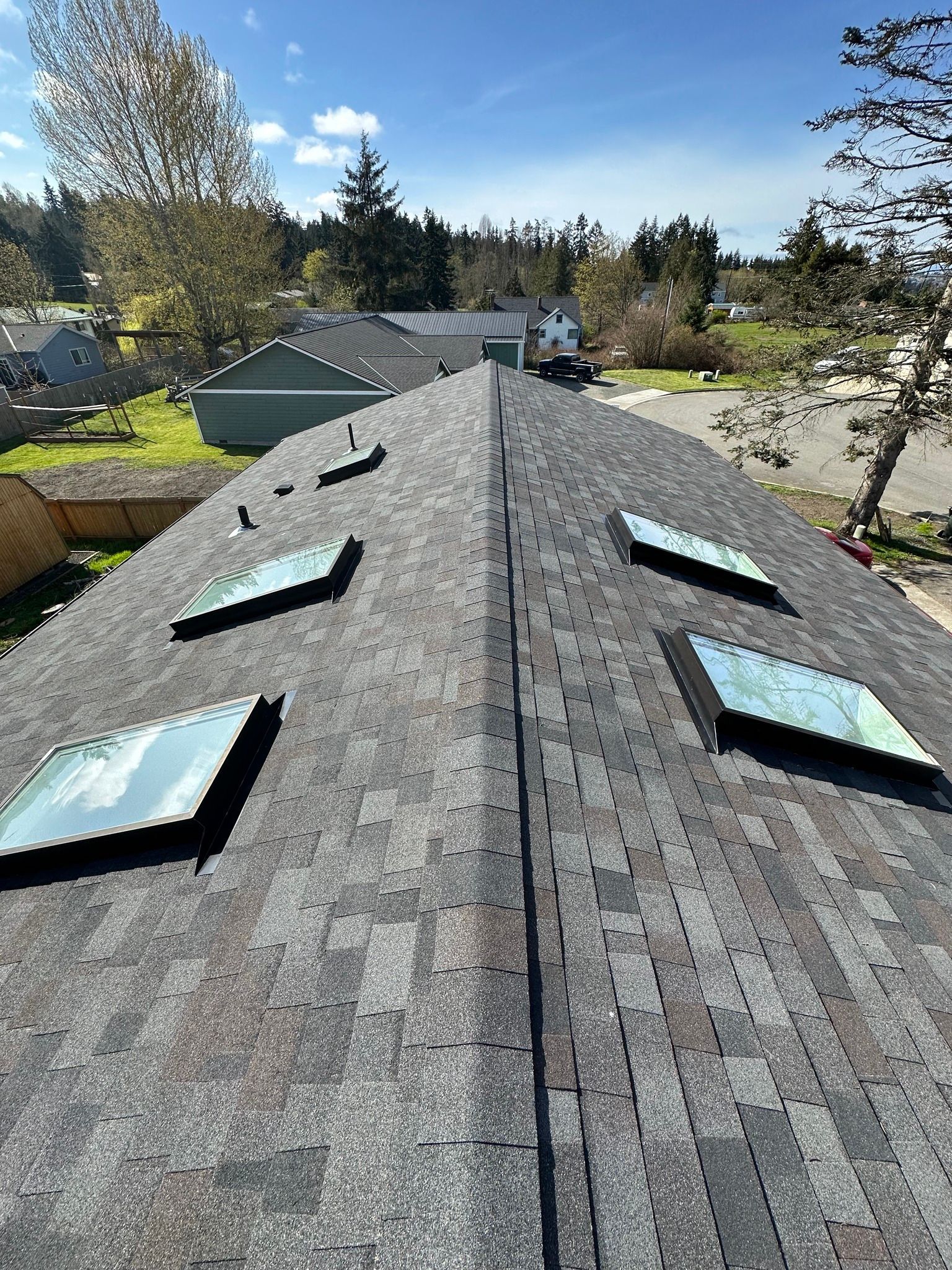 View of a roof with skylights and asphalt shingles under a blue sky, trees, and buildings in the background.