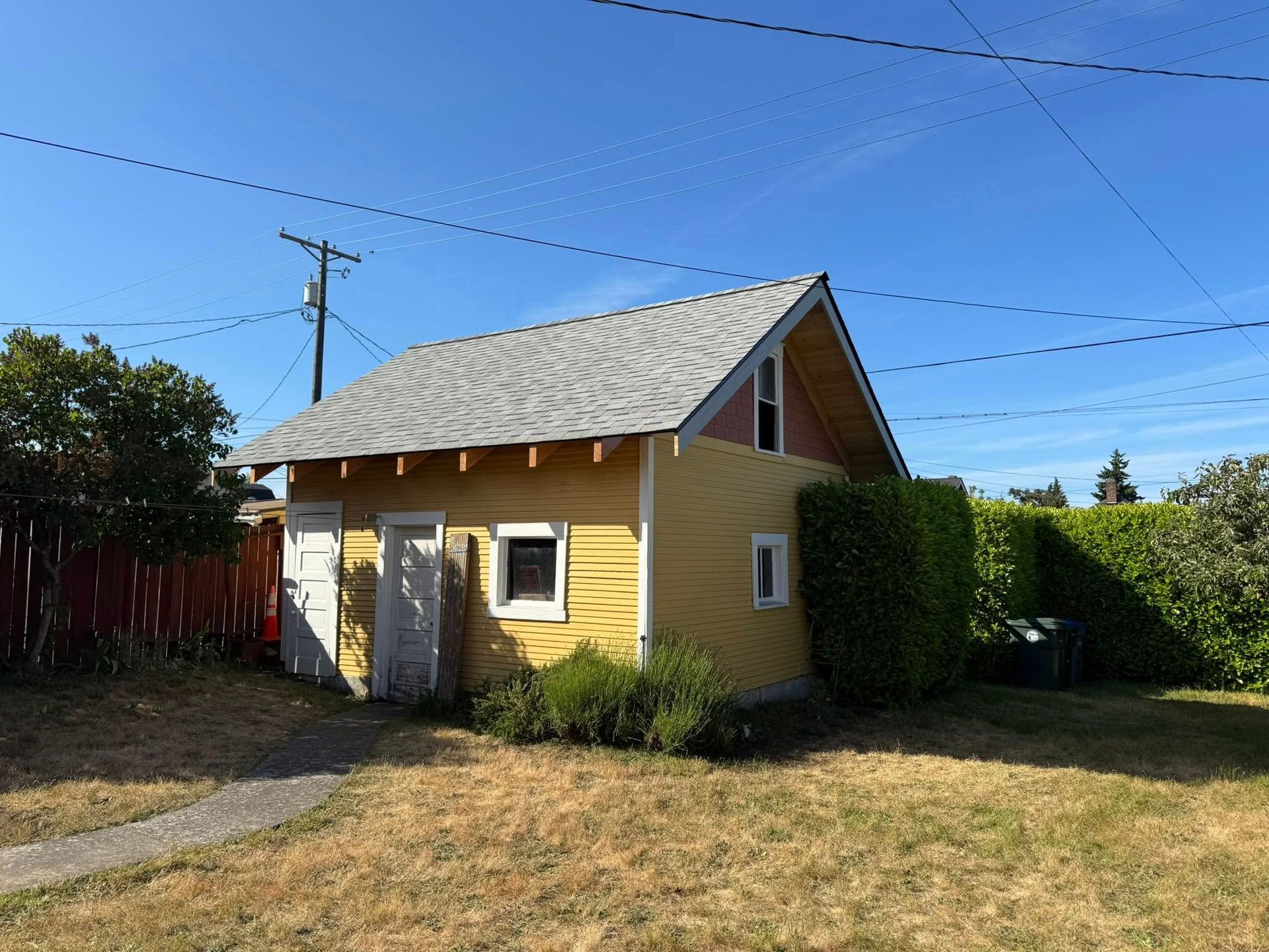 Yellow shed with gray roof, small windows, and hedge. Blue sky background.