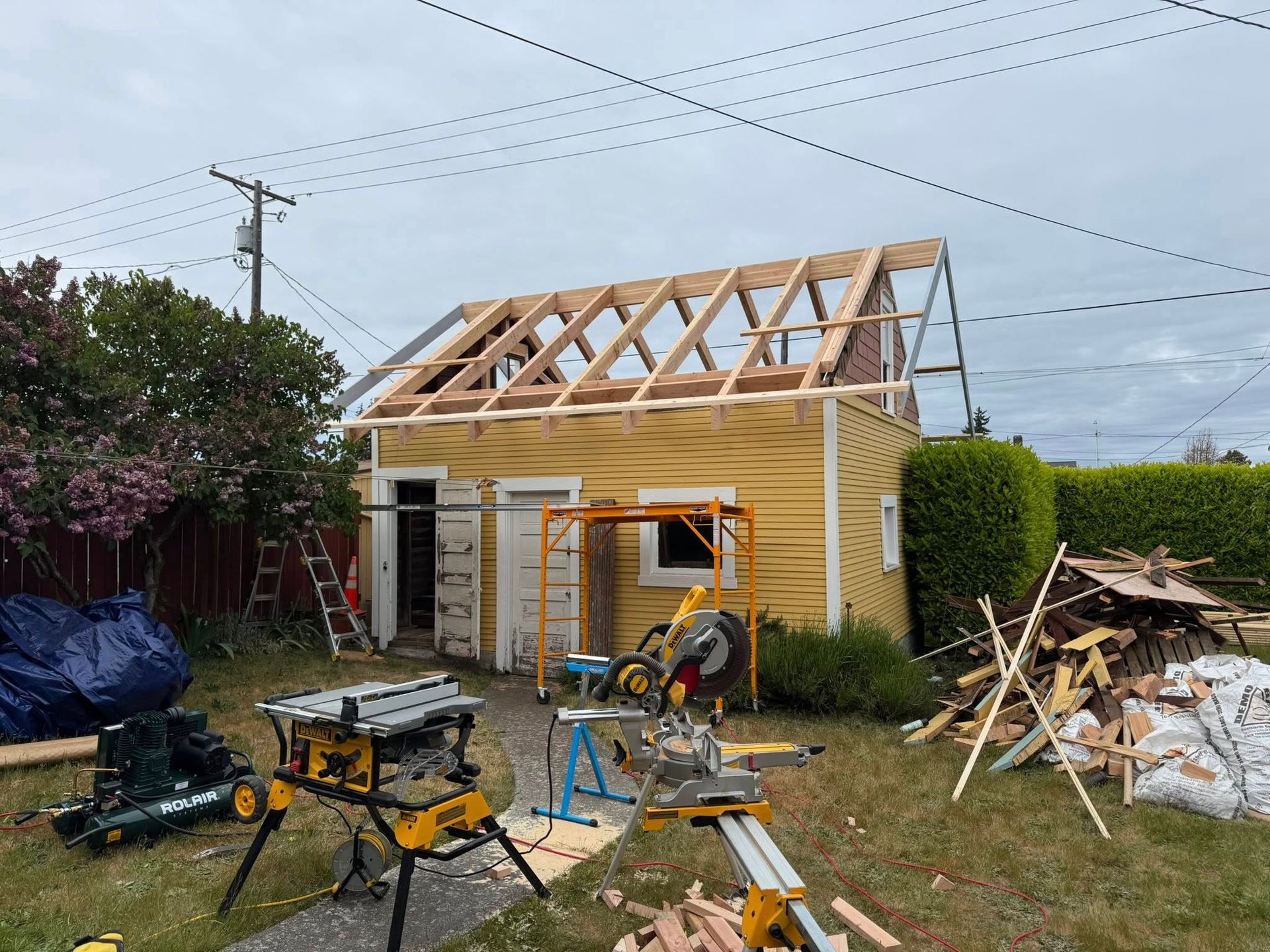 Construction of a shed; yellow exterior, wooden roof frame. Tools and lumber in yard, cloudy sky.