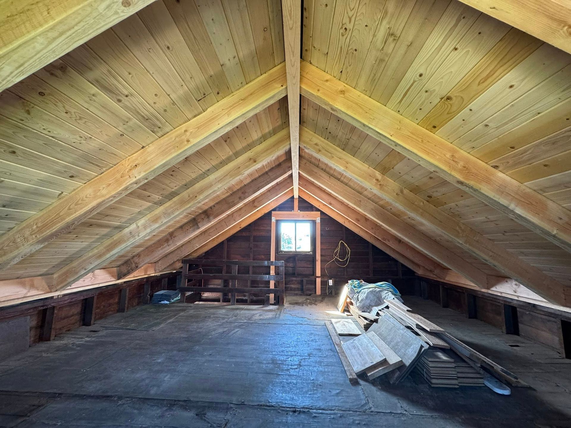 Attic space with wooden beams, ceiling, and floor; a small window in the background.